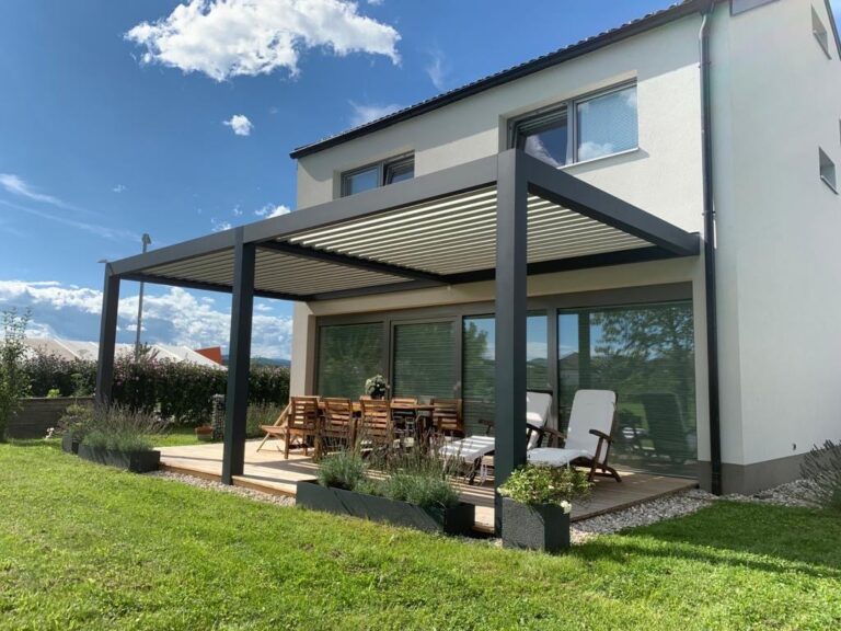 Dark-framed louvred pergola shading a dining table and loungers beside a white house